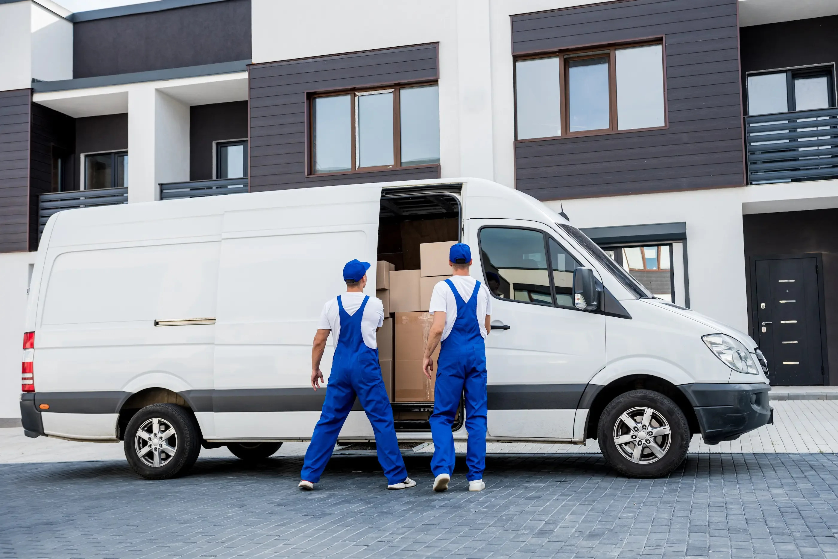 Two movers in blue uniforms are loading cardboard boxes into a white moving van.