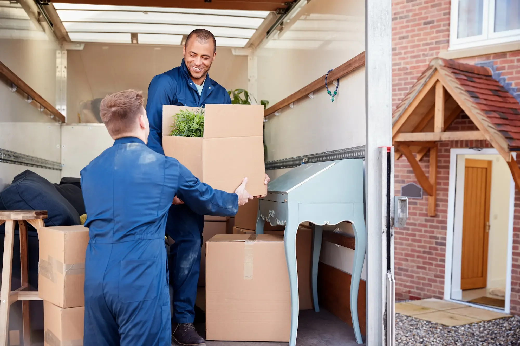 Two movers load boxes and furniture into a removal van outside a brick house